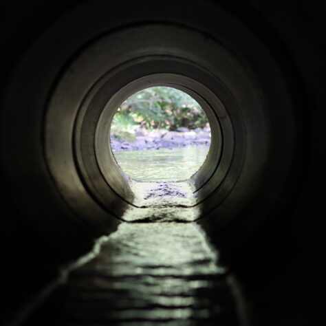 View through a round concrete tunnel carrying a stream beneath a road