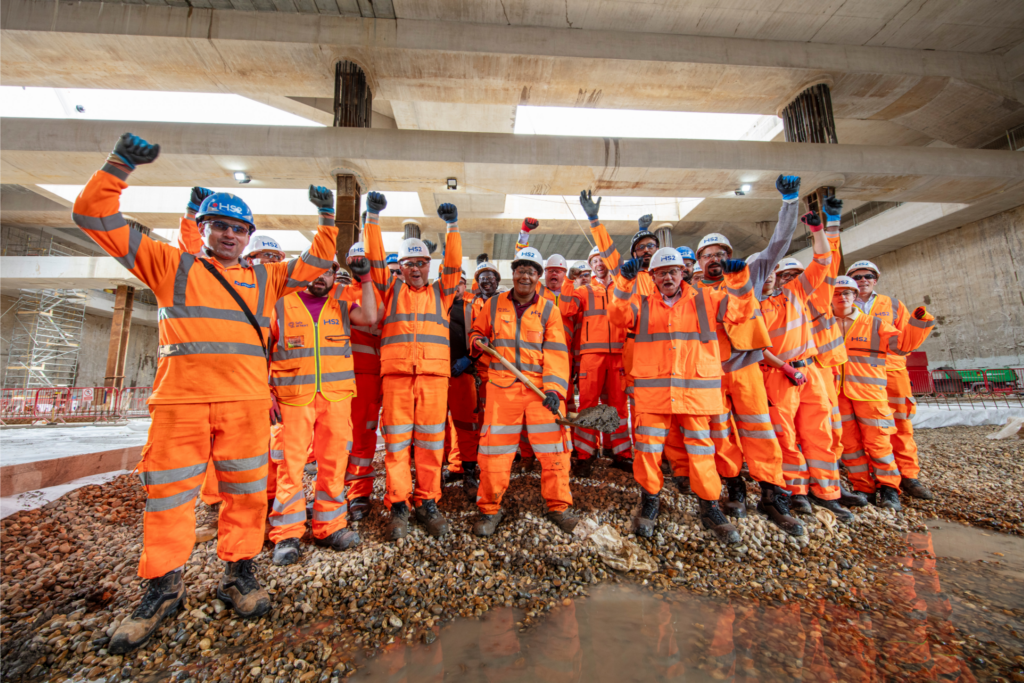 The team at Old Oak Common celebrates the final excavation of the Old Oak Common box. They are stood in a group in the construction area of the project, all wearing orange high-vis PPE. Some of the group have their fists raised in the air and they are all cheering and smiling.