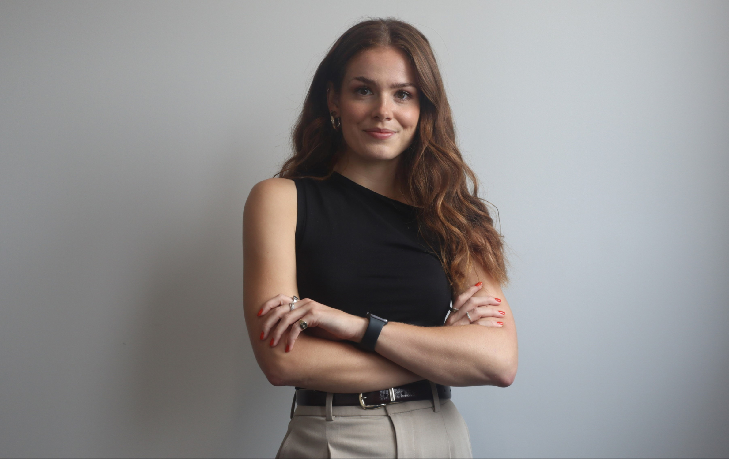 A professional headshot photo of Riona Ainge (Sustainability Lead at SYSTRA). Riona is wearing a black top, beige trousers and has her arms crossed. She is looking at the camera smiling. The background is grey.