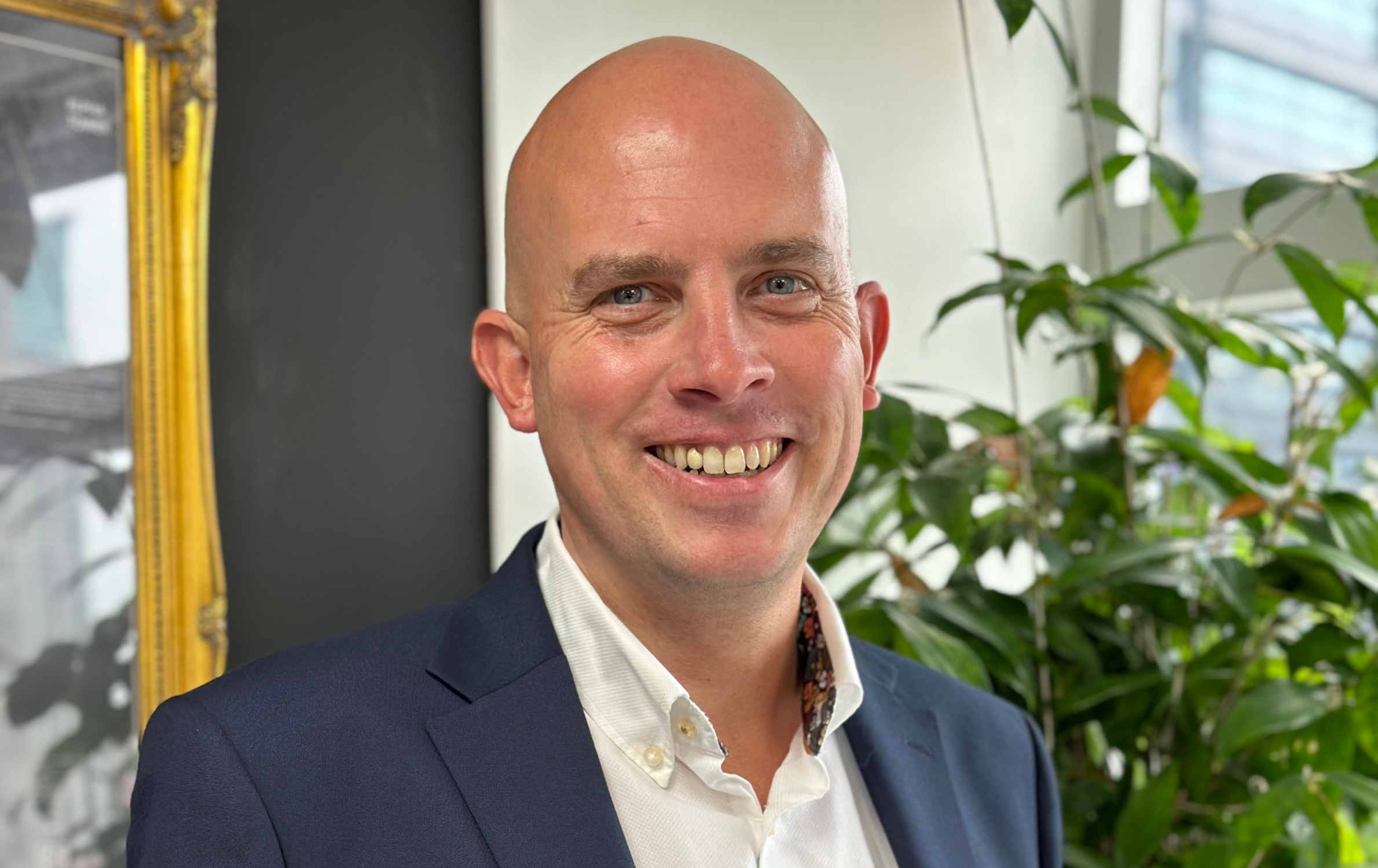 A headshot image of James Easty. James is wearing a suit jacket and is smiling at the camera. He is stood in one of the SYSTRA offices - behind him is a plant and a mirror on the wall.