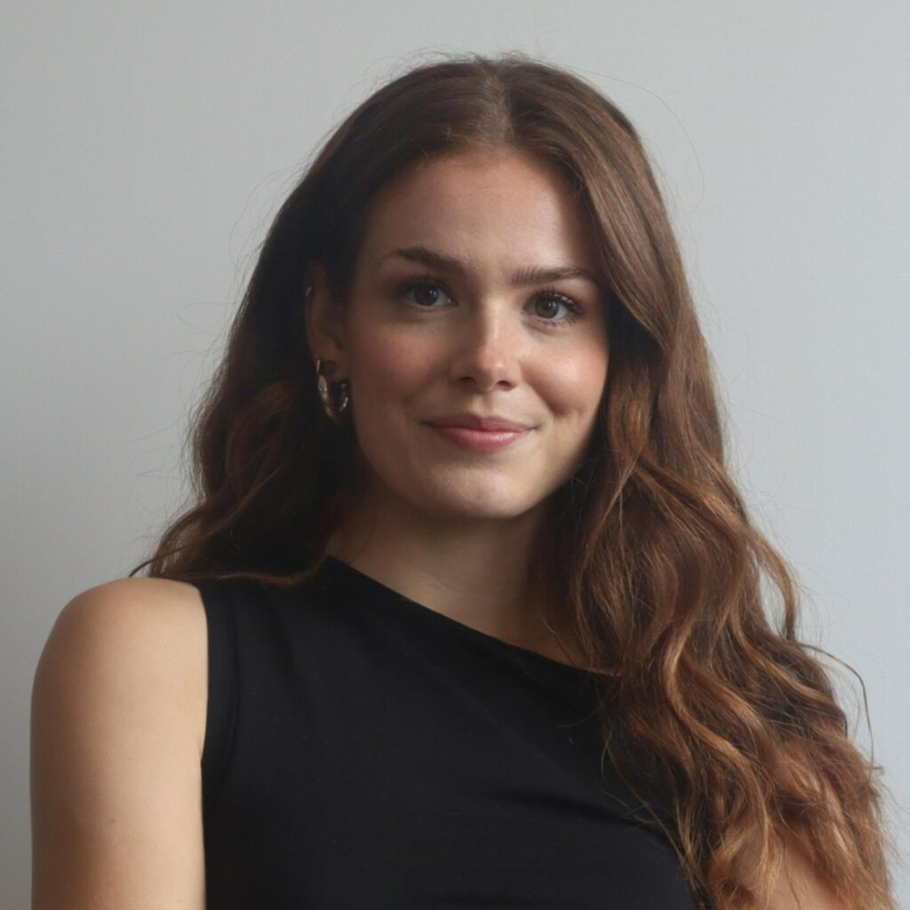 A professional headshot photo of Riona Ainge (Sustainability Lead at SYSTRA). Riona is wearing a black top and is looking at the camera smiling. The background is grey.