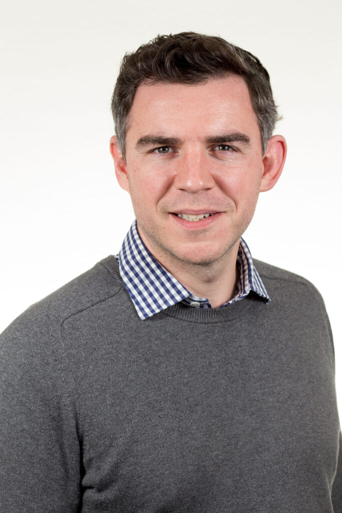A headshot image of Malcolm Calvert. Malcolm is wearing a grey jumper and is smiling at the camera. The backdrop is white.