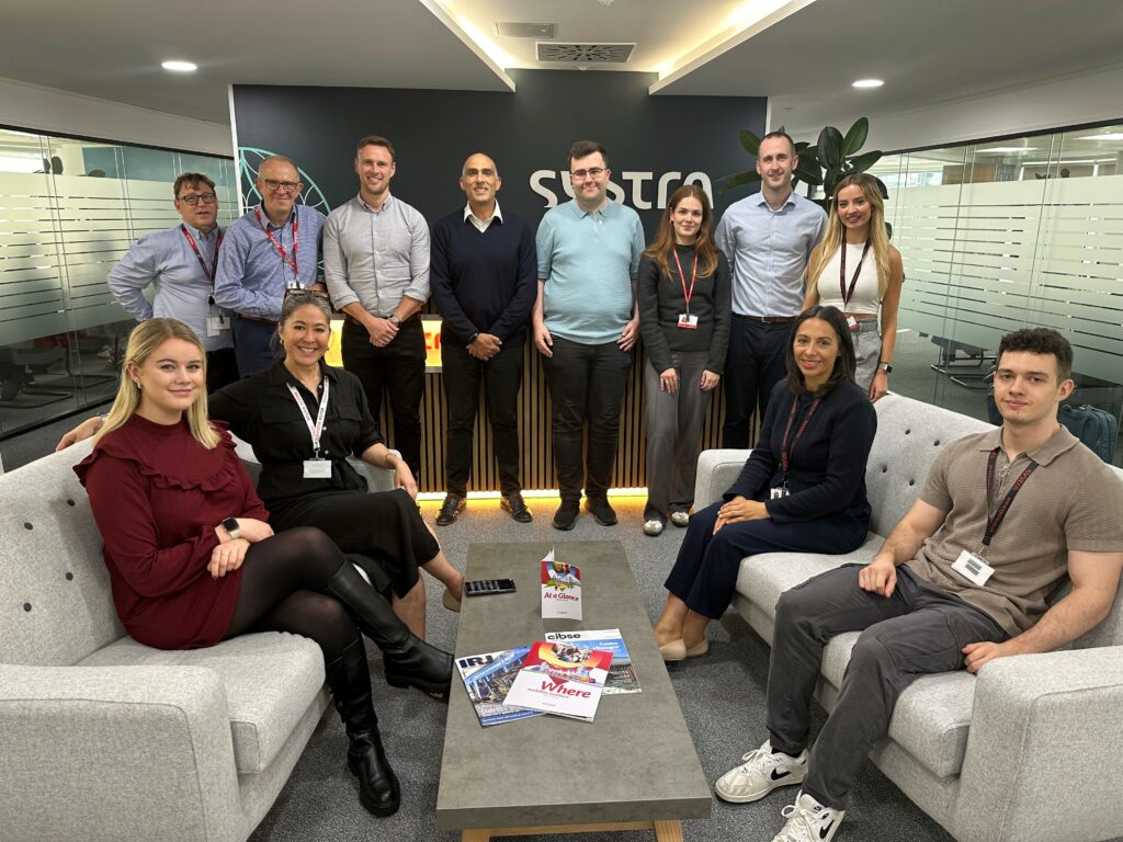 SYSTRA colleagues who took part in the carbon literacy training gather round for a photo in the office. Some are sat either side on 2 sofas, and the rest are stood in a line at the back. They are all smiling at the camera.