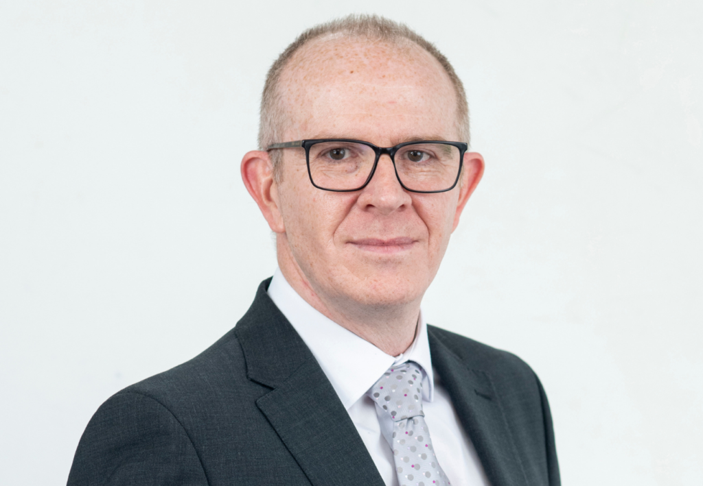 A professional headshot photo of David White (Director of Health, Safety, Security & Sustainability at SYSTRA). David is wearing a smart suit and tie, glasses and is looking at the camera smiling. The background is white.