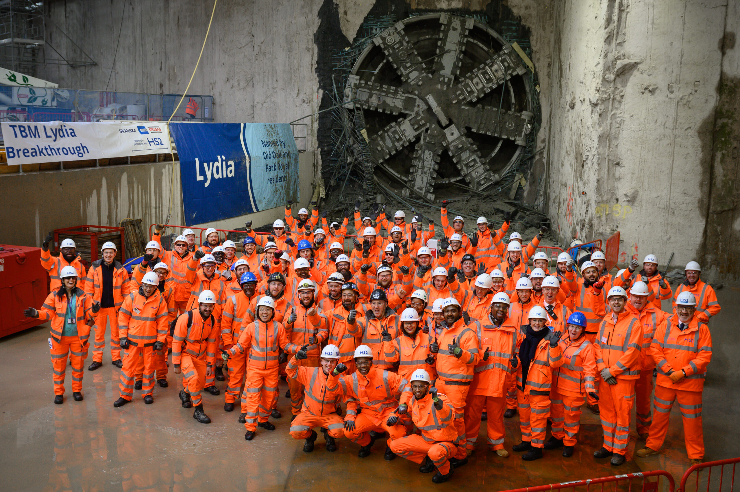 A photo of the big Old Oak Common team celebrating in front of the 'Lydia' tunnel boring machine which broke the into the station box. They are all in bright orange his vis PPE.
