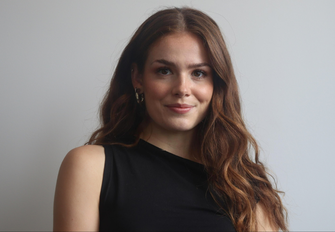 A professional headshot photo of Riona Ainge (Sustainability Lead at SYSTRA). Riona is wearing a black top and is looking at the camera smiling. The background is grey.