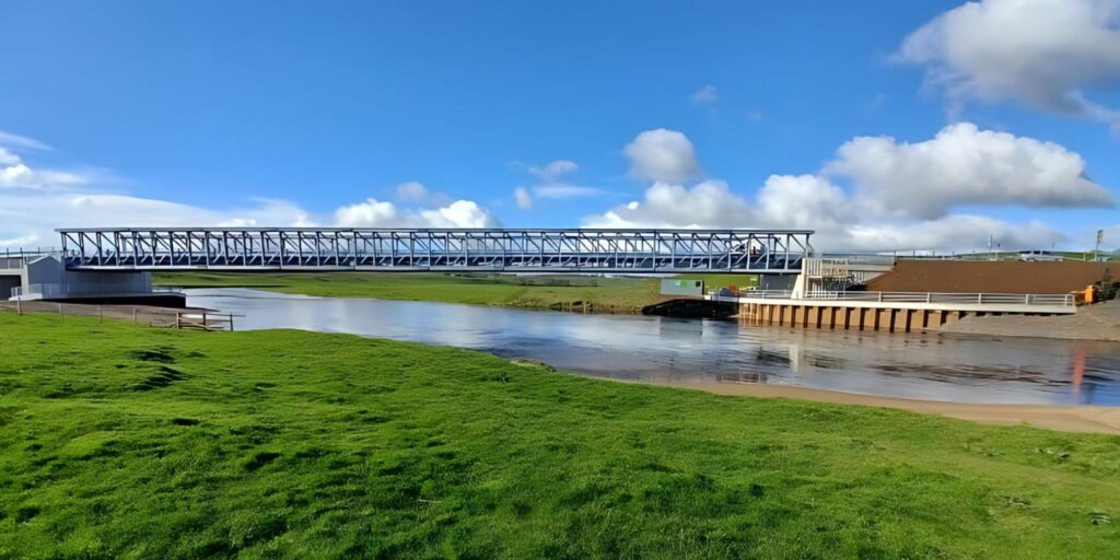 A picturesque photo of the new permanent River Clyde crossing in South Lanarkshire. Around the river is luscious green grass and the sky is blue with bright white clouds. It is a sunny day.