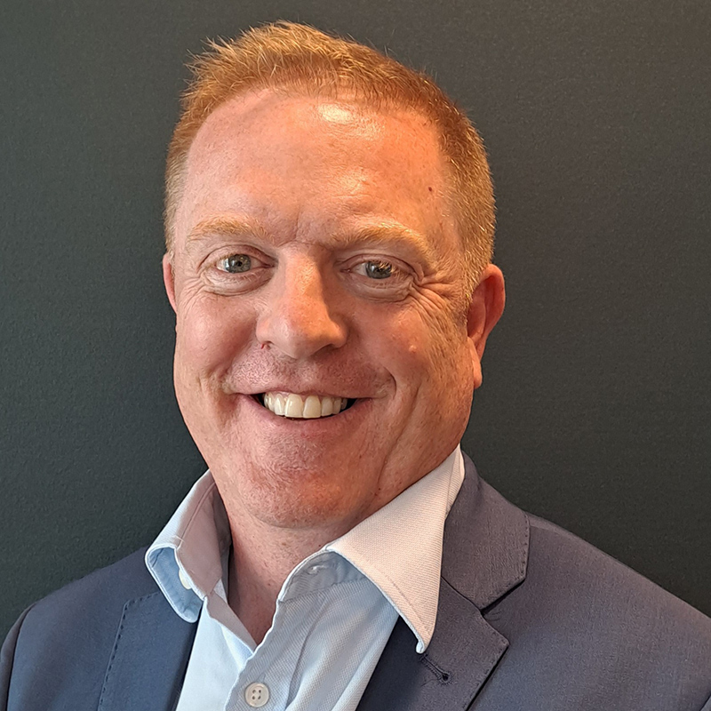 A close up headshot photo of Andrew Dugdale. Andrew is wearing a shirt and suit jacket and is smiling at the camera.