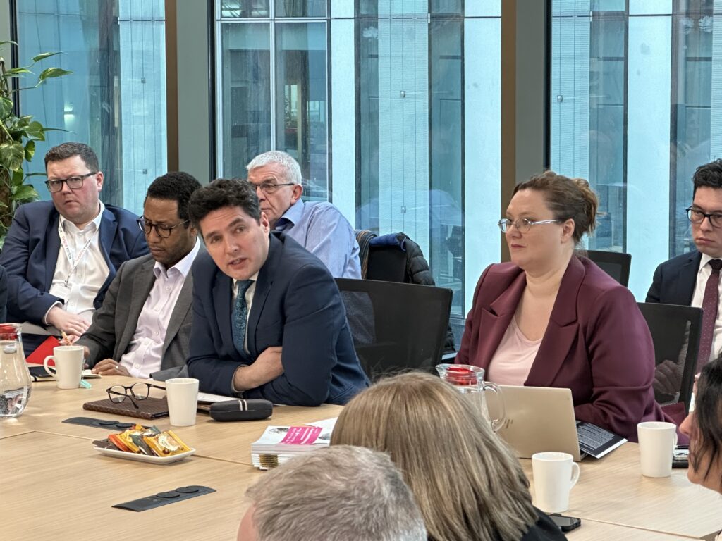 A photo of Huw Merriman at the RIA meeting in the SYSTRA Manchester office. Huw is sat with his armed folded and is leaning forward on a large table. Other meeting attendees are sat next to and behind him. In the background are other offices buildings seen through a large window.