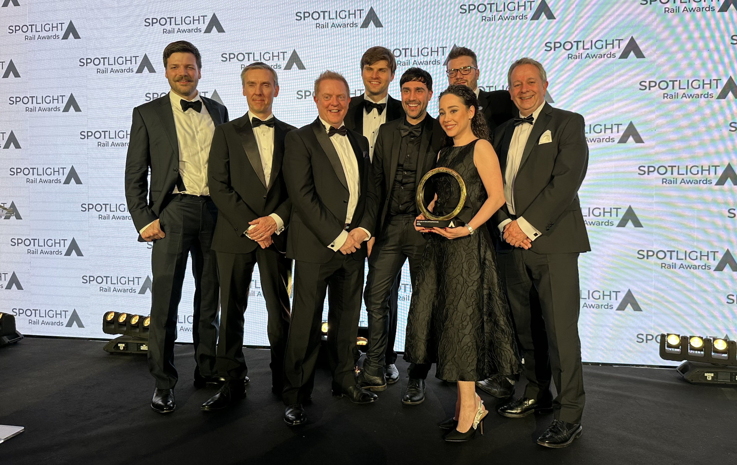 A photo of award winners at the SPOTLIGHT Rail Awards 2025. They are pictured standing in a group holding their trophy. Behind them is a wall with the event logo repeated across it.