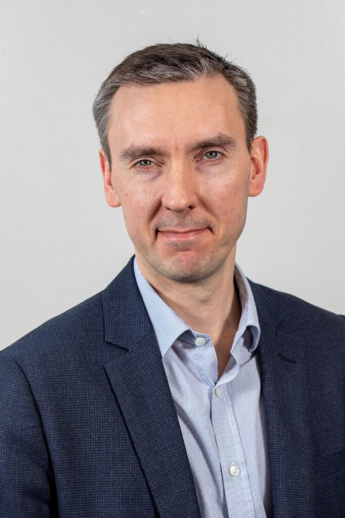 Headshot photo of Rob Dale. He is wearing a shirt and suit jacket and is smiling at the camera. The background is plain.