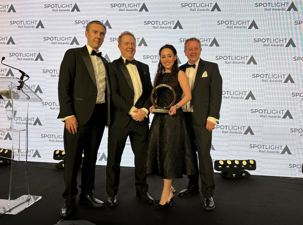A photo of 4 award winners at the SPOTLIGHT Rail Awards 2025. They are pictured standing in a group holding their trophy. Behind them is a wall with the event logo repeated across it.