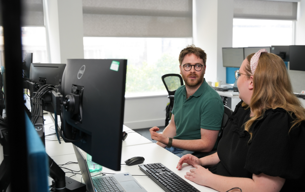 Image of two SYSTRA colleagues collaborating at a work desk in a SYSTRA office. They are facing each other and are talking. The photo is taken from the side and there are two large desktop computer screens at the desk.