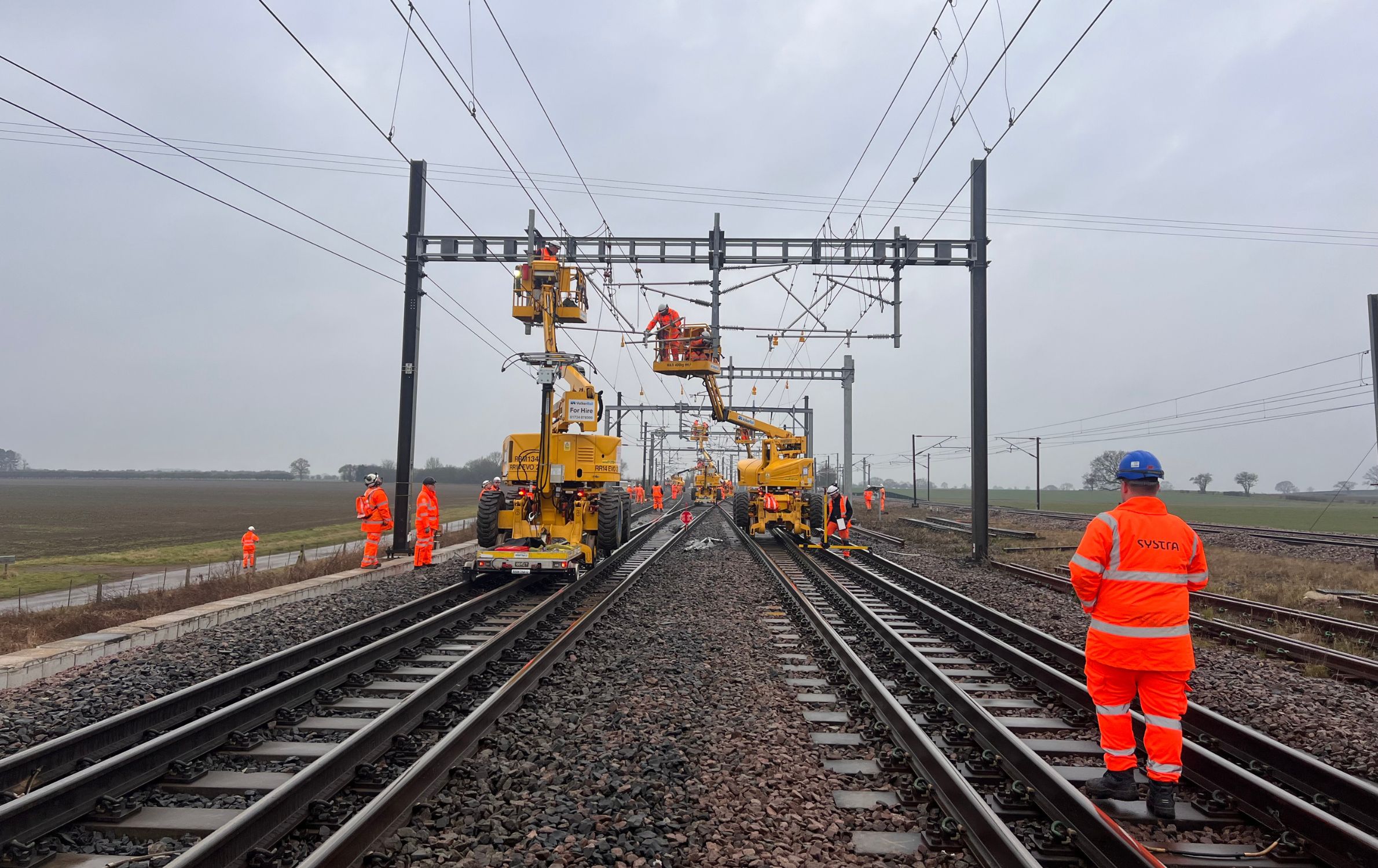 A photo of the TRU Colton Junction. The photo is taken on the tracks as they stretch out underneath OLE wires. Construction vehicles and people wearing SYSTRA branded bright orange PPE are pictured on the tracks.