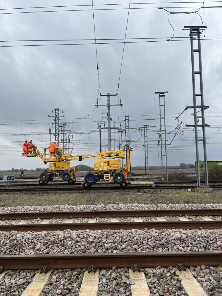 A photo of the TRU Colton Junction. The photo is taken from the side of the tracks underneath OLE wires. Construction vehicles, with people wearing SYSTRA branded bright orange PPE on them, are pictured on the tracks.