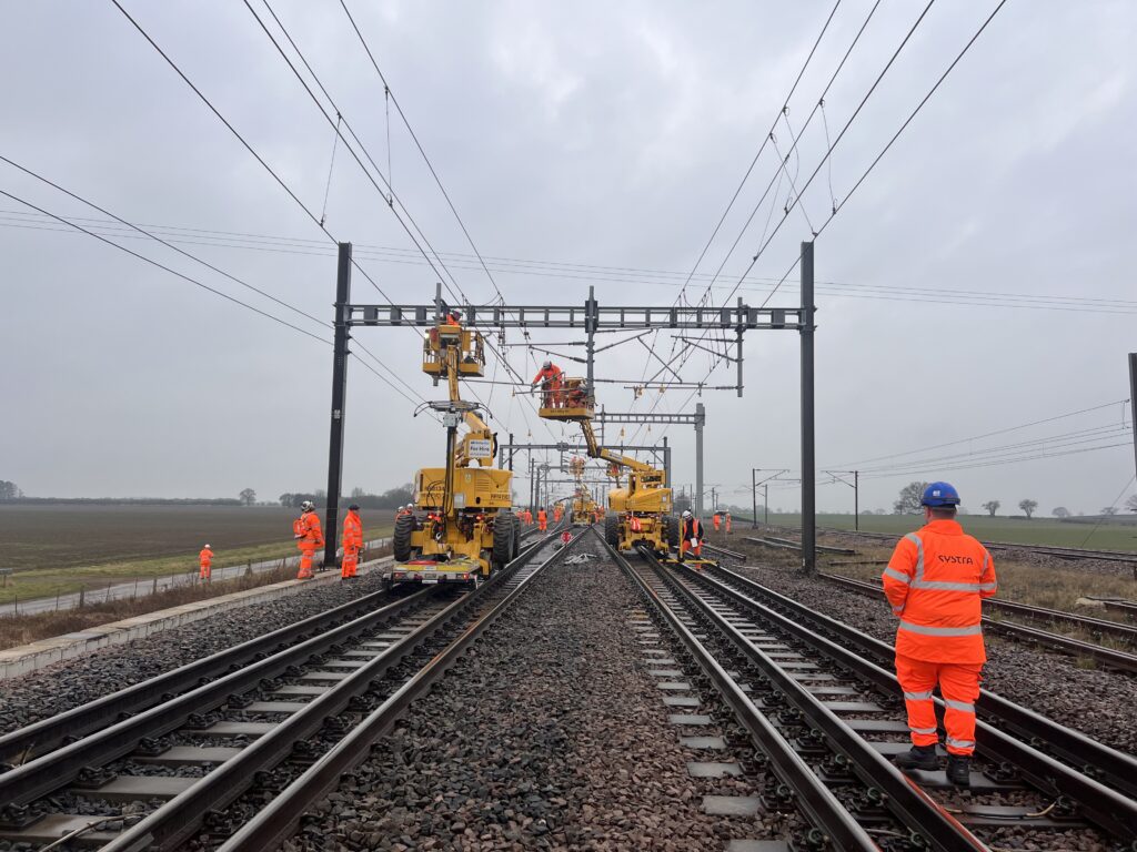 A photo of the TRU Colton Junction. The photo is taken on the tracks as they stretch out underneath OLE wires. Construction vehicles and people wearing SYSTRA branded bright orange PPE are pictured on the tracks.