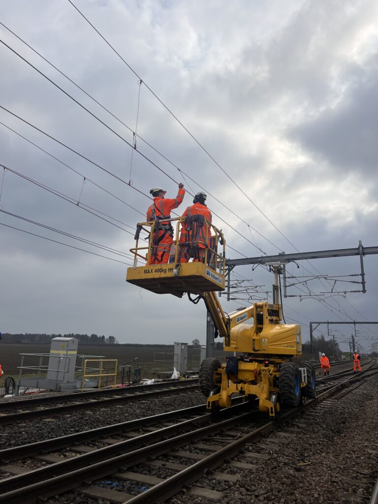 A photo of the TRU Colton Junction. The photo is taken from an angle at the side of the tracks underneath OLE wires. A construction vehicle, with people wearing SYSTRA branded bright orange PPE on it, is pictured on the tracks.