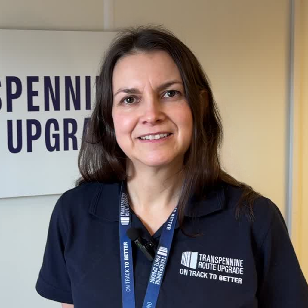 A photo of Steph Whiteley at the TRU offices. She has long dark brown hair, is wearing a navy blue polo top with the TRU logo on it, a lighter blue TRU lanyard, and is smiling at the camera.