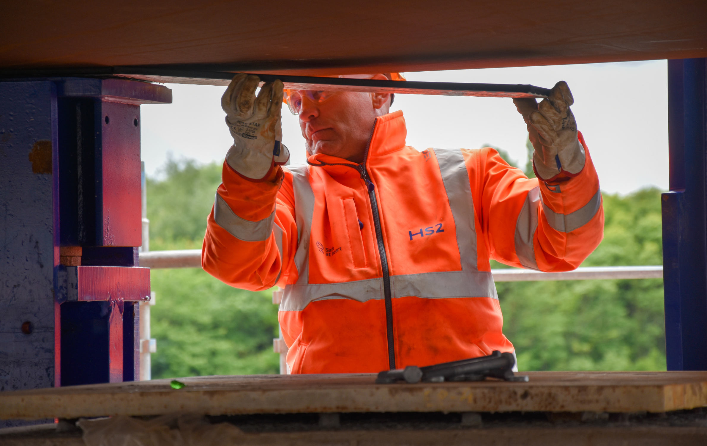 A construction worker wearing bright orange PPE is pictured inserting non-stick pads under the deck beam of the M6 South viaduct deck on the HS2 project. The photo is taken through a 'window' of the deck beam. Behind the construction worker are lots of blurred trees.