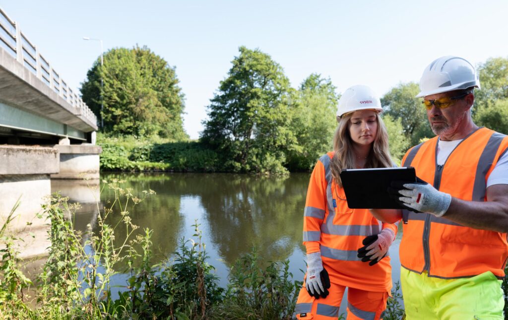 SYSTRA environment specialists safely using a tablet near water and a bridge