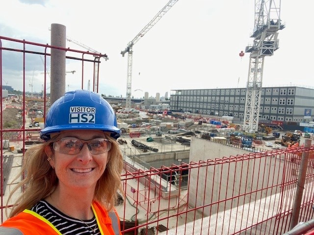 Jennifer Jefferies is pictured at the HS2 Old Oak Common site. She is taking a selfie and smiling at the camera. She is wearing a blue hard hat and a high-vis jacket. Behind her is the Old Oak Common site in progress including large cranes.
