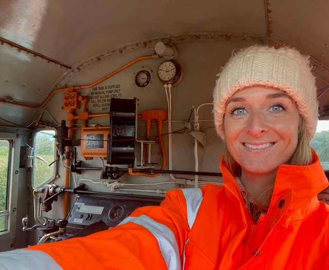 Jennifer Jefferies is pictured with a high-vis jacket and wooley hat on, in the front of a train. She is smiling and taking a selfie.