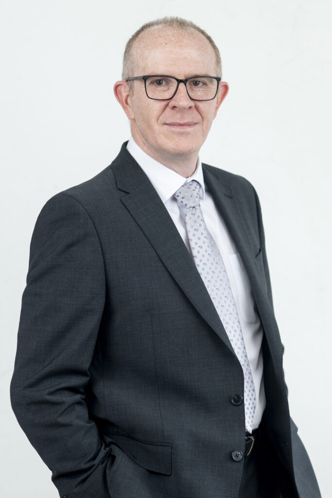 A professional headshot photo of David White (Director of Health, Safety, Security & Sustainability at SYSTRA). David is wearing a smart suit and tie, glasses and is looking at the camera smiling. The background is white.