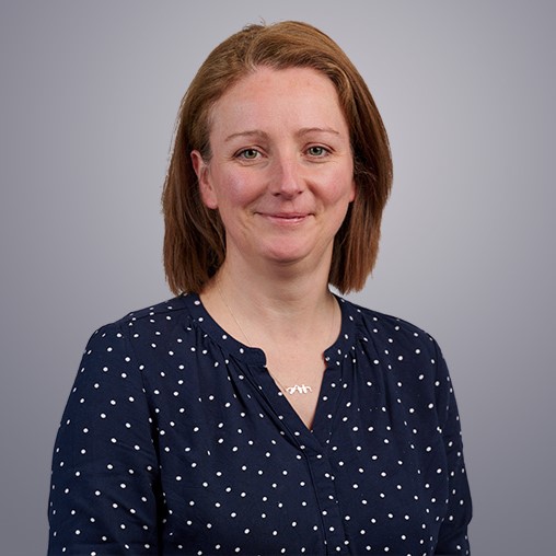 A headshot photo of Amy Sykes. She is wearing a dark long-sleeved top, has short red hair and is smiling at the camera. The background is grey.