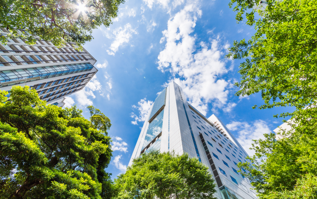 A photo taken looking up at two high rise buildings and blue sky and clouds. Surrounding the buildings are lots of green luscious trees.