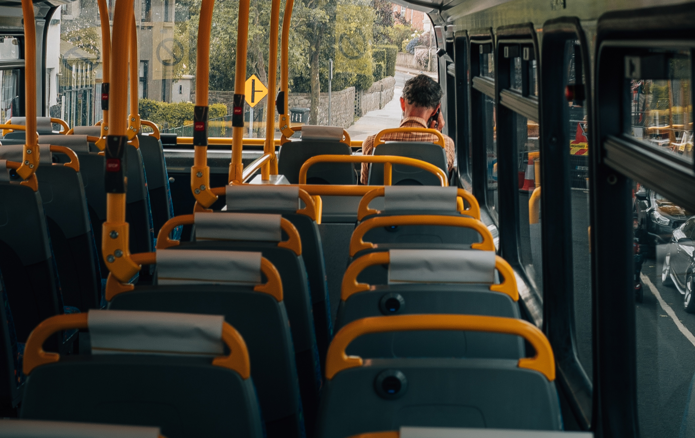Photo of the inside of a bus taken from behind lots of empty seats. At the front a man is sat with a checked orange shirt on and is on the phone.