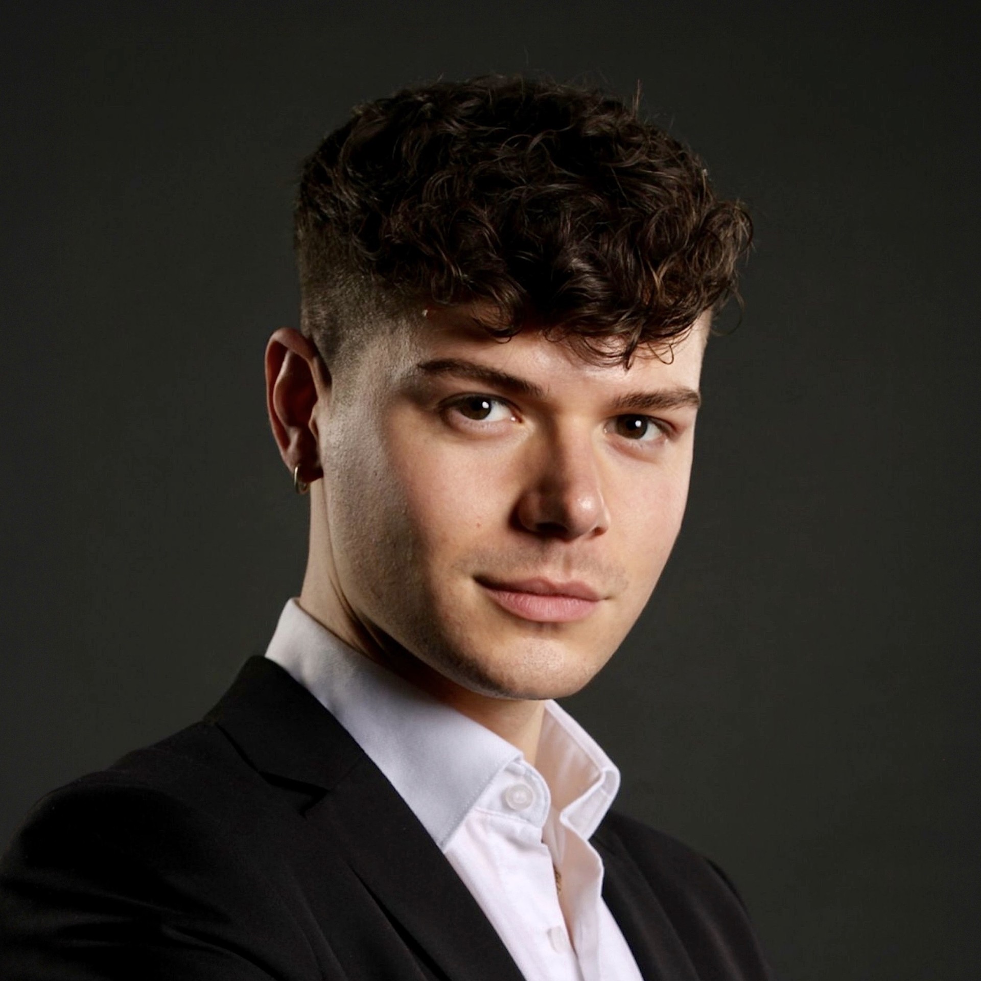 A headshot photo of Joe Thompson, PlanBEE Rail apprentice. Joe is wearing a white shirt and a black suit jacket, and is looking at the camera. The background is dark.