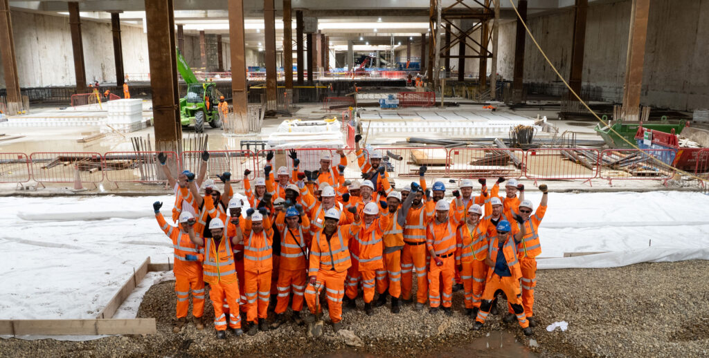 The construction team at HS2's Old Oak Common station celebrate the final excavation of the box. They are stood in a group on the construction site with their hands in the air smiling. They are all wearing bright orange PPE. The photo is taken from above with a drone.