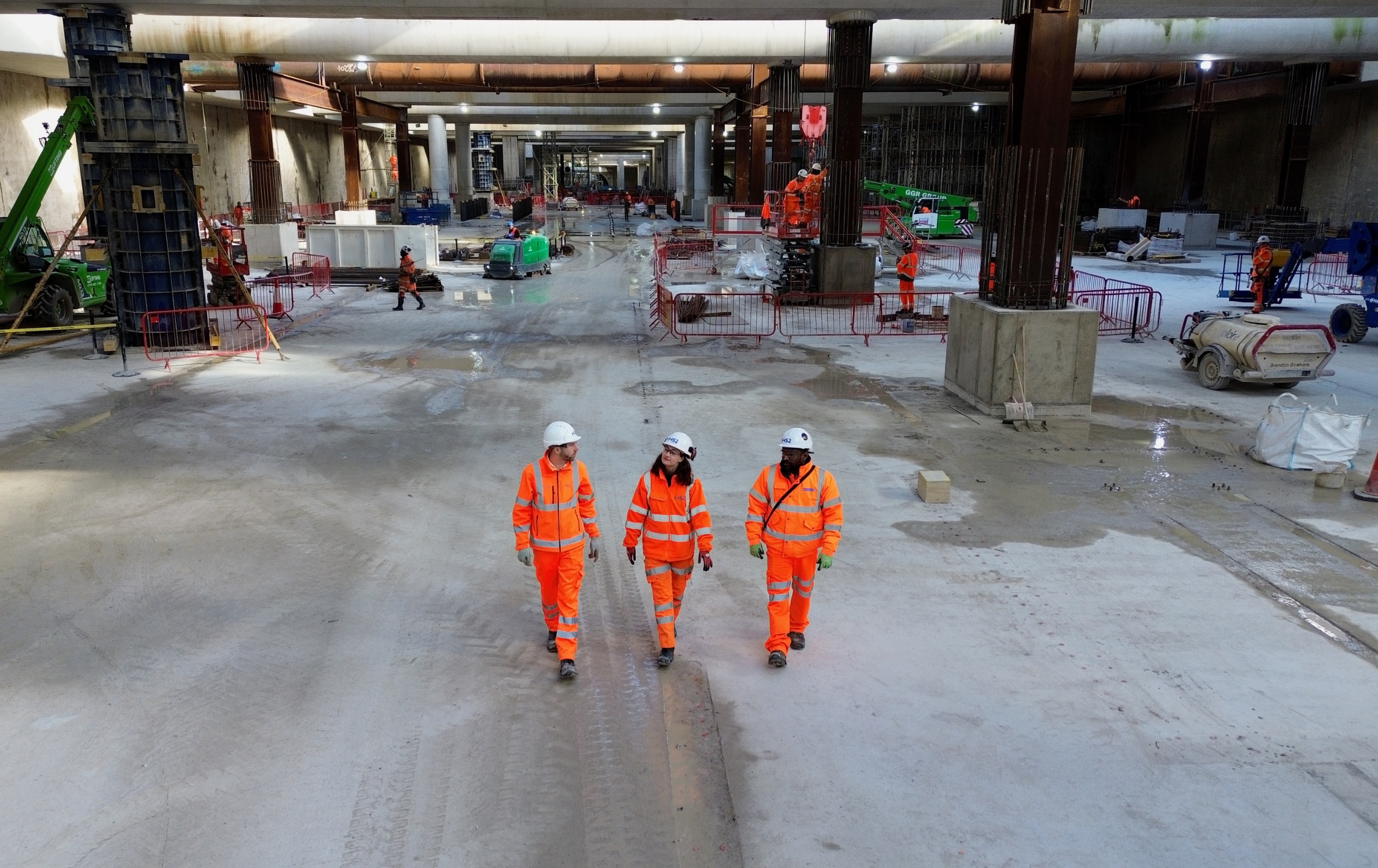 Three people wearing bright orange PPE walk through the middle of the HS2 Old Oak Common construction site where the foundations have been completed. The photo is taken from above.