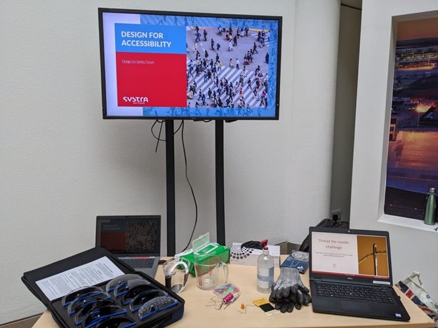 A photo of a small meeting room. There is a TV screen showing a SYSTRA branded presentation that is titled 'Design for accessibility'. The round table in front is full of items such as a needle and thread, plastic bottle, gloves and laptop.