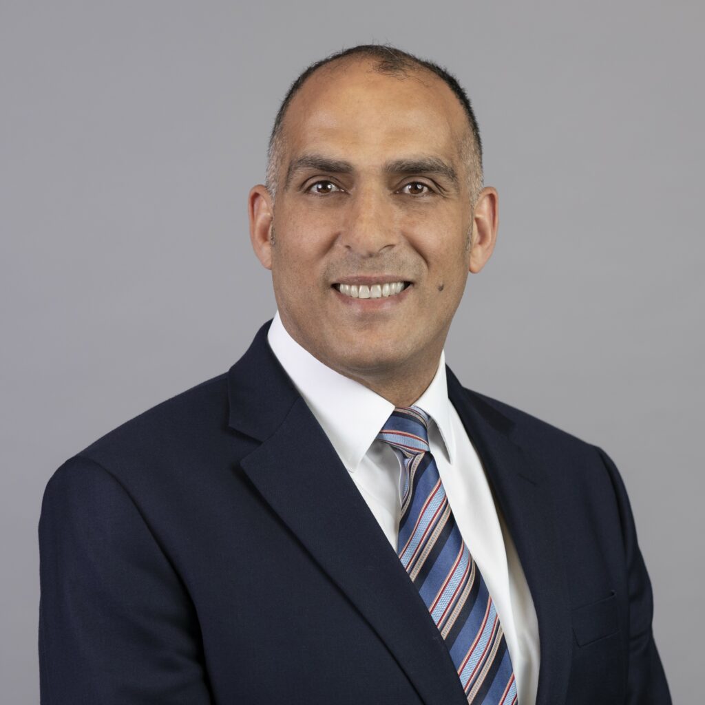 Headshot photo of Nizar Awad. Nizar is wearing a white shirt, black suit jacket and stripey tie. He is smiling at the camera and there is a grey background.