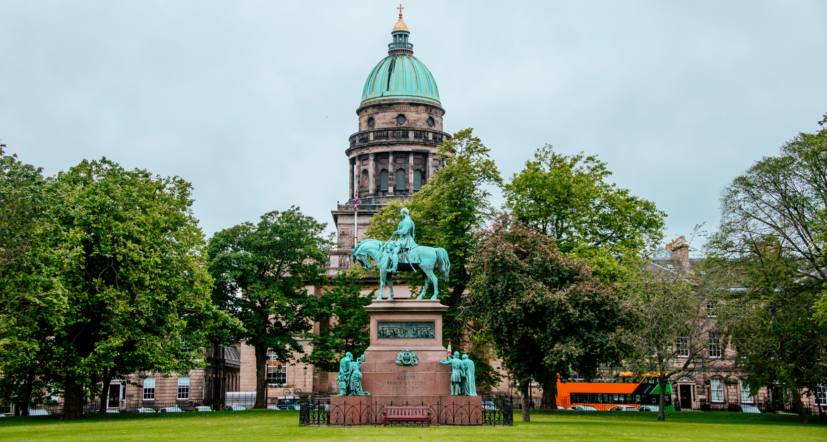 A photo of a park in Edinburgh. There is a grand looking building with a green dome in the background. In the middle of the shot is a statue of a man on a horse. It says 'Albert Prince Consort' at the bottom of the statue. The park is surrounded by trees.