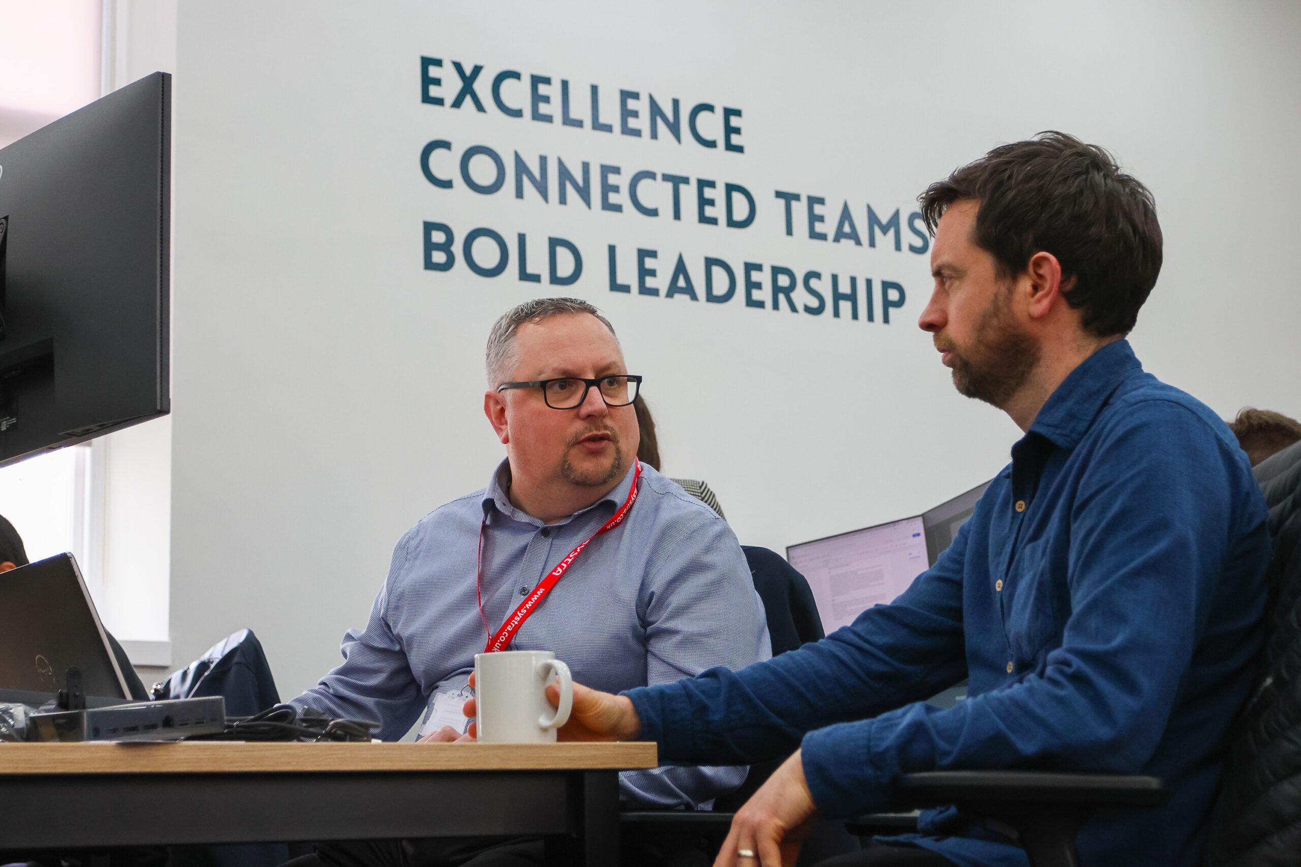 Image of two SYSTRA colleagues collaborating at a work desk in a SYSTRA office. They are talking to each other and there is a computer desktop screen on the left-hand side of the photo. Behind them the SYSTRA values are on the wall. It reads 'Excellence', 'Connected Teams' and 'Bold Leadership'.