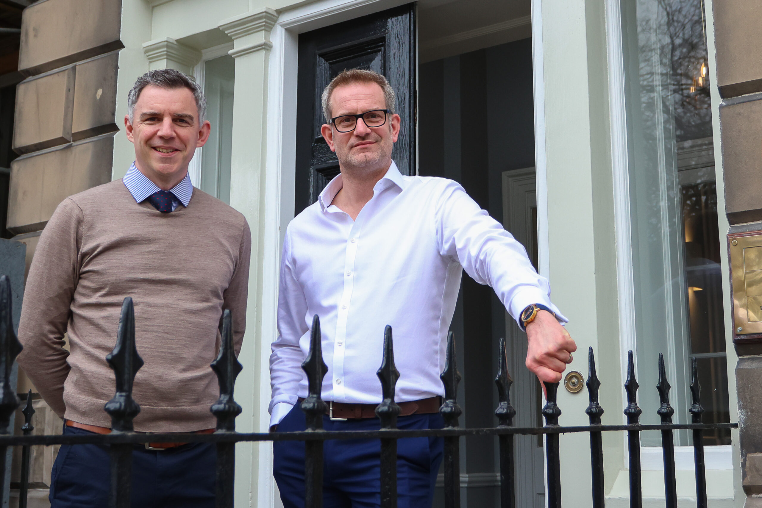 Photo of Keith McGillivray standing outside the entrance to the SYSTRA Edinburgh office next to a colleague. He is wearing white shirt and black suit trousers, and has his hand on a black metal fence. His colleague is wearing black suit trousers and a brown jumper. They are both smiling at the camera.
