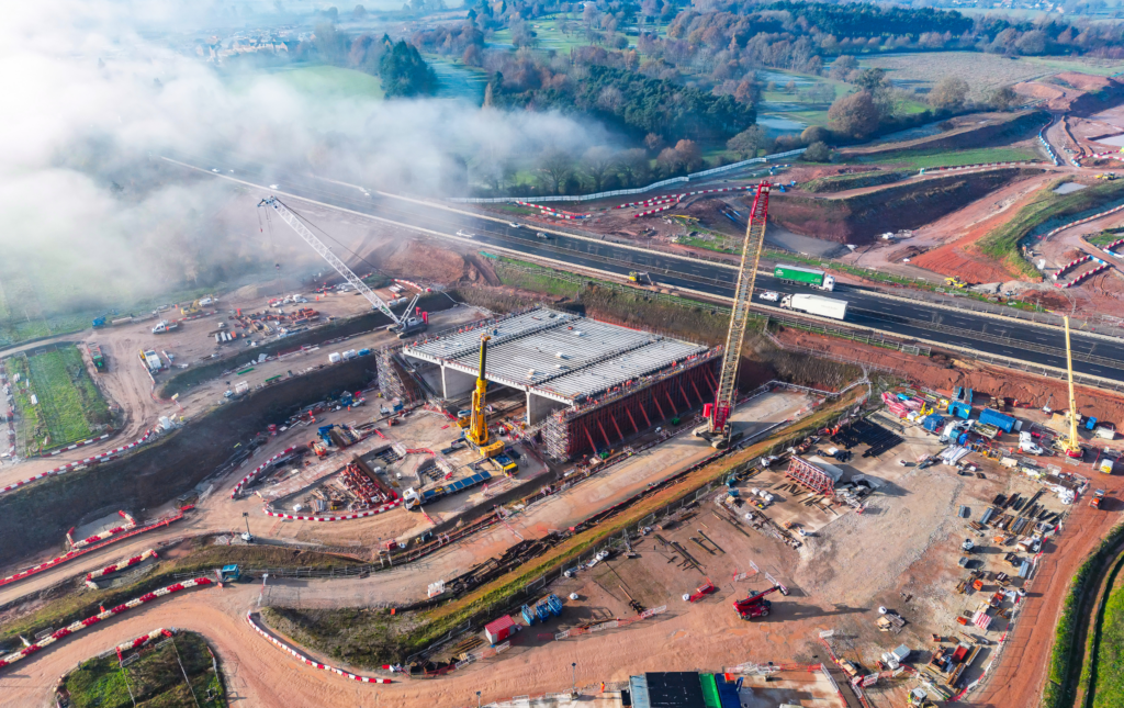HS2 construction site at the A46 Kenilworth bypass. The photo shows the bridge's box structure next to the highway. There are a number of cranes and the construction site looks busy. In the background are lots of trees and fields.
