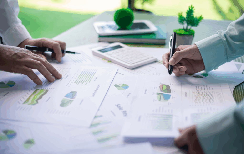 Printed sustainability reports spread out across a table. On either side are the hands of people in business attire. They are both holding pens.