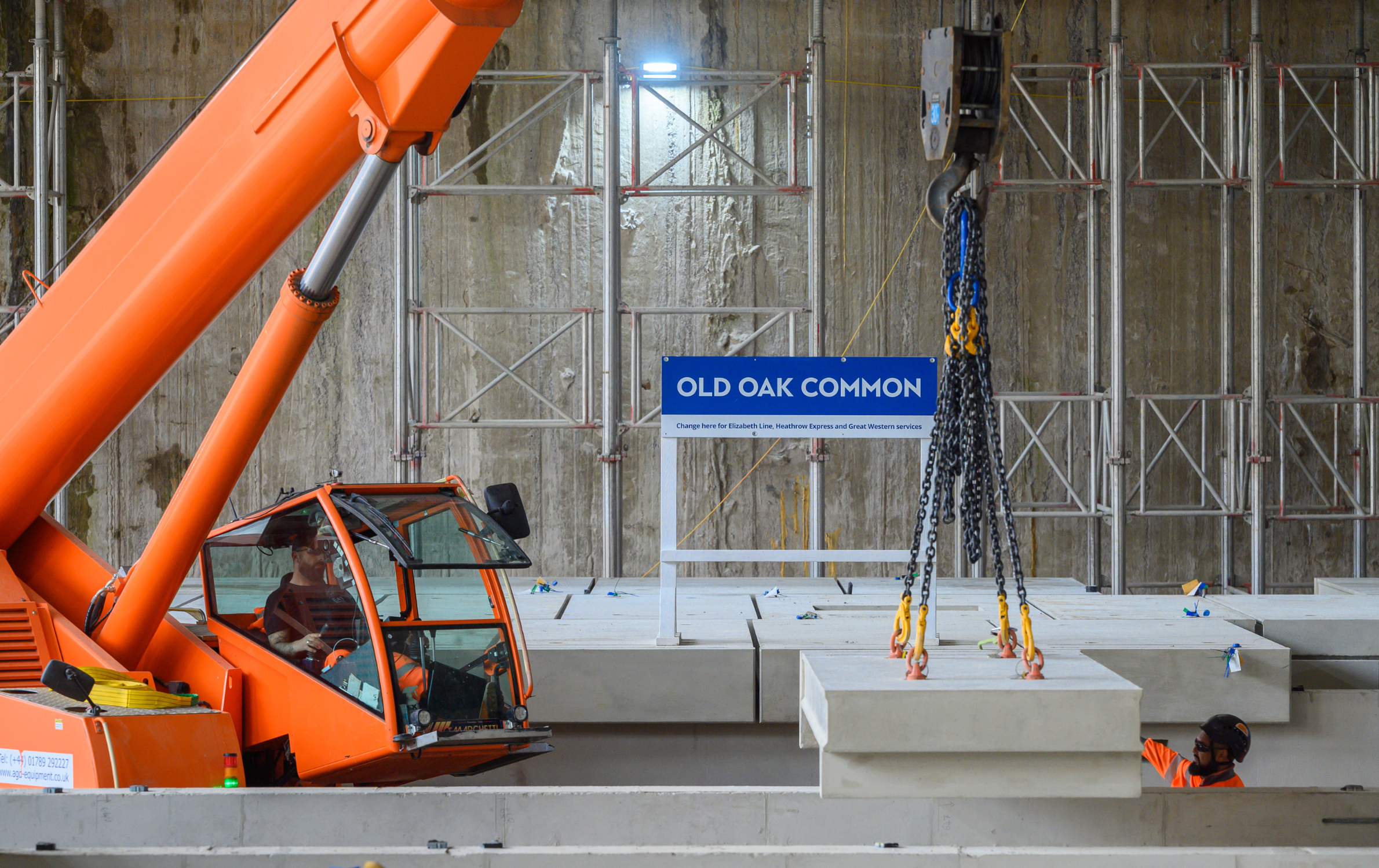 A photograph of two workers wearing PPE at the HS2 Old Oak Common station construction site where the first platforms are being installed. One of them is sat inside a bright orange digger, and the other is stood where the rail tracks run. They are both below platform level. On the platform there is a bright blue sign that says Old Oak Common.